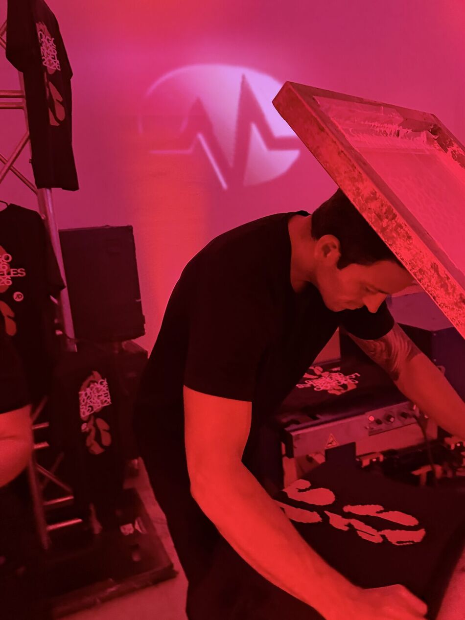 A printer lowering an inked screen onto a shirt at the press during an evening event